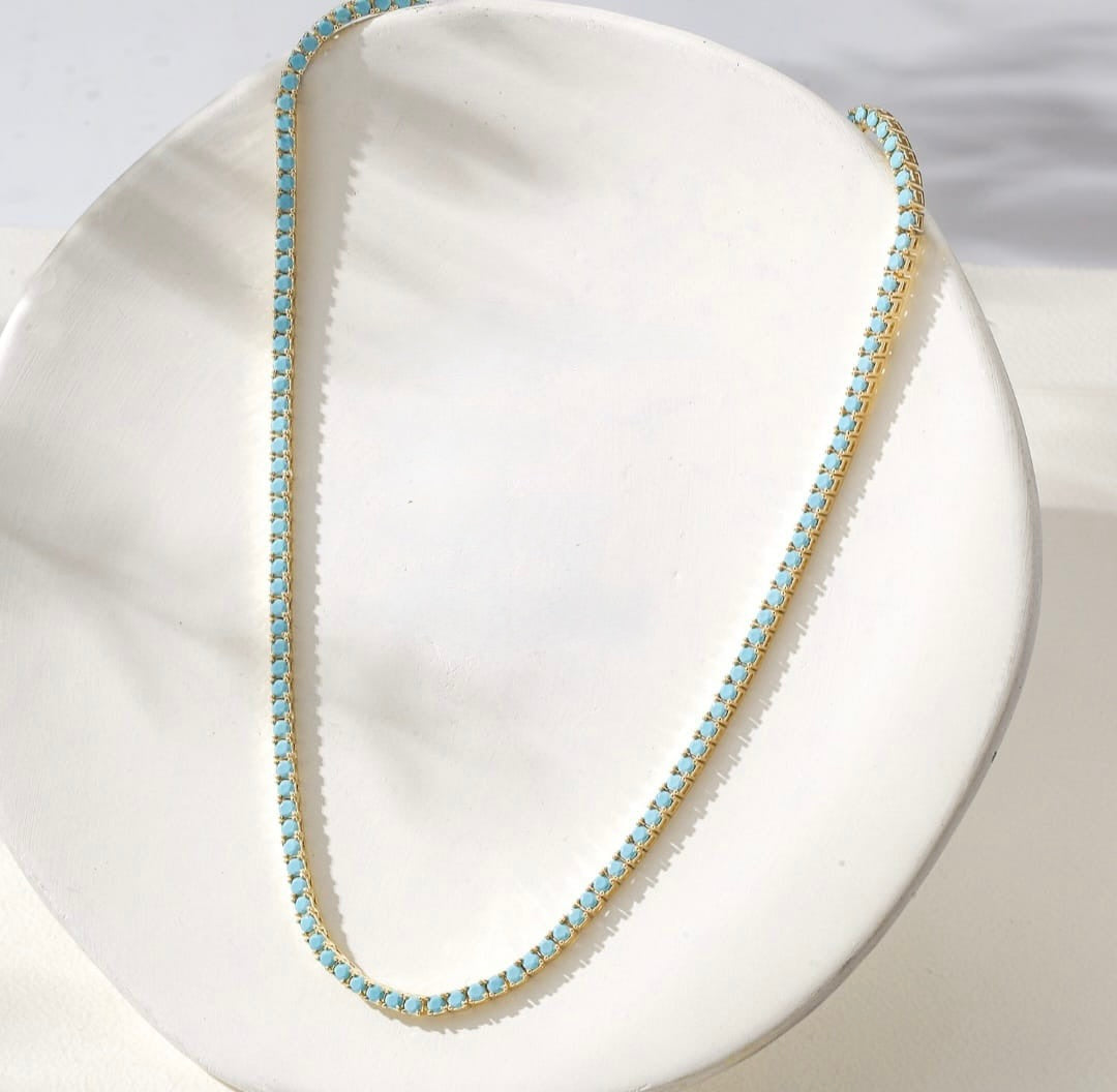 A delicate gold chain necklace with small blue turquoise stones, draped over a white ceramic display dish.