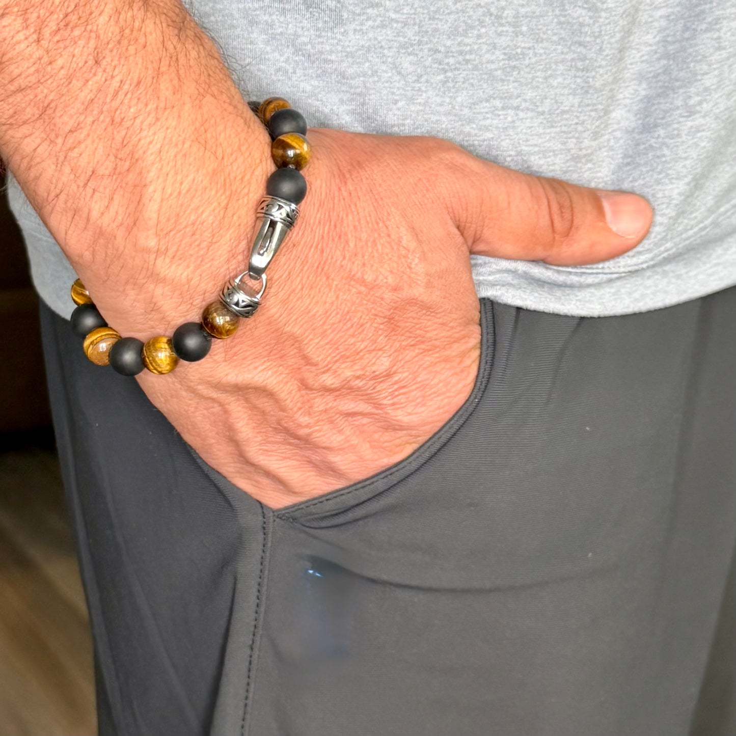 A close-up of a man's hand resting in his pocket, wearing a beaded bracelet with matte black, polished brown, and tiger's eye beads, and a silver-toned metal clasp.