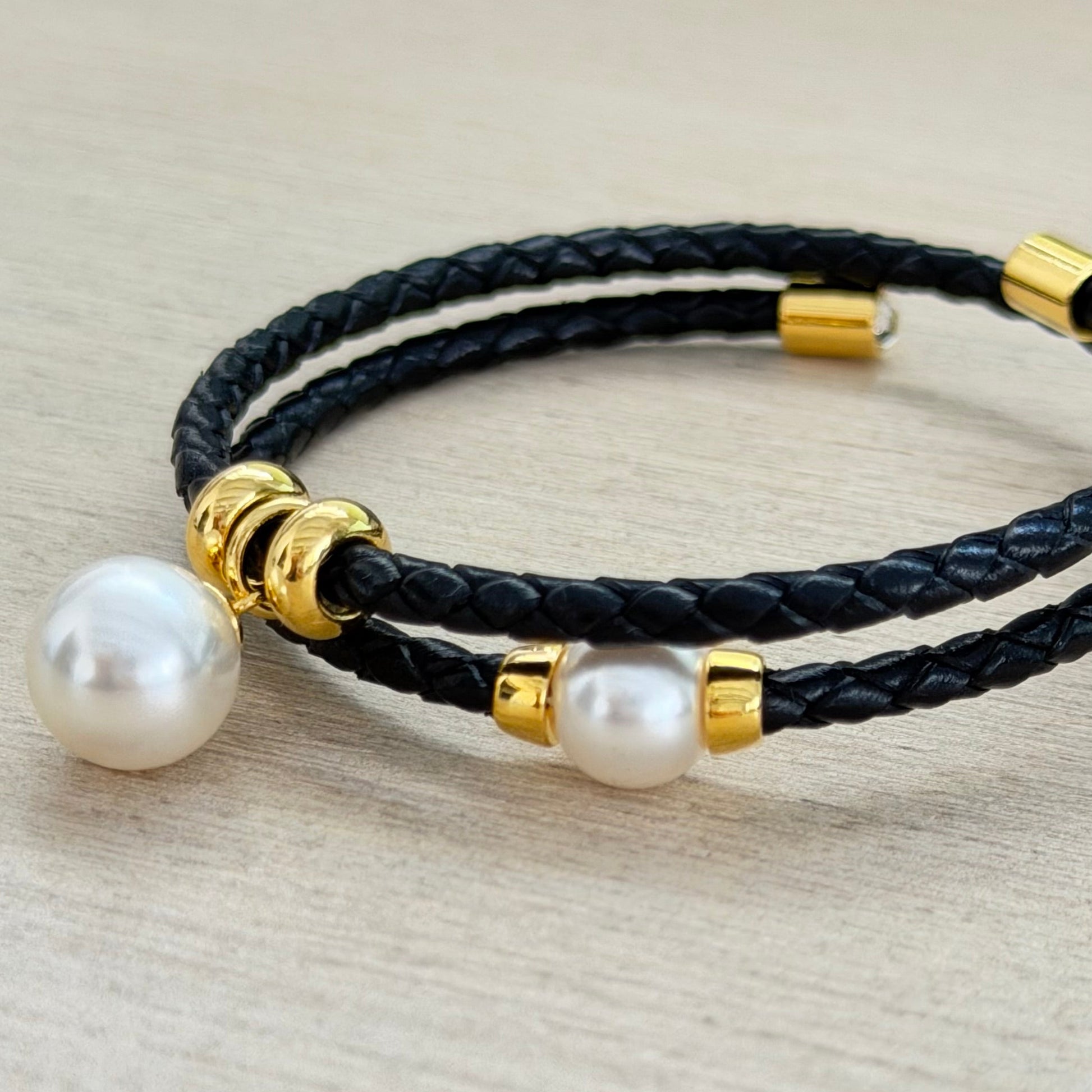 Black braided leather bracelet with gold accents and pearls on a light gray background