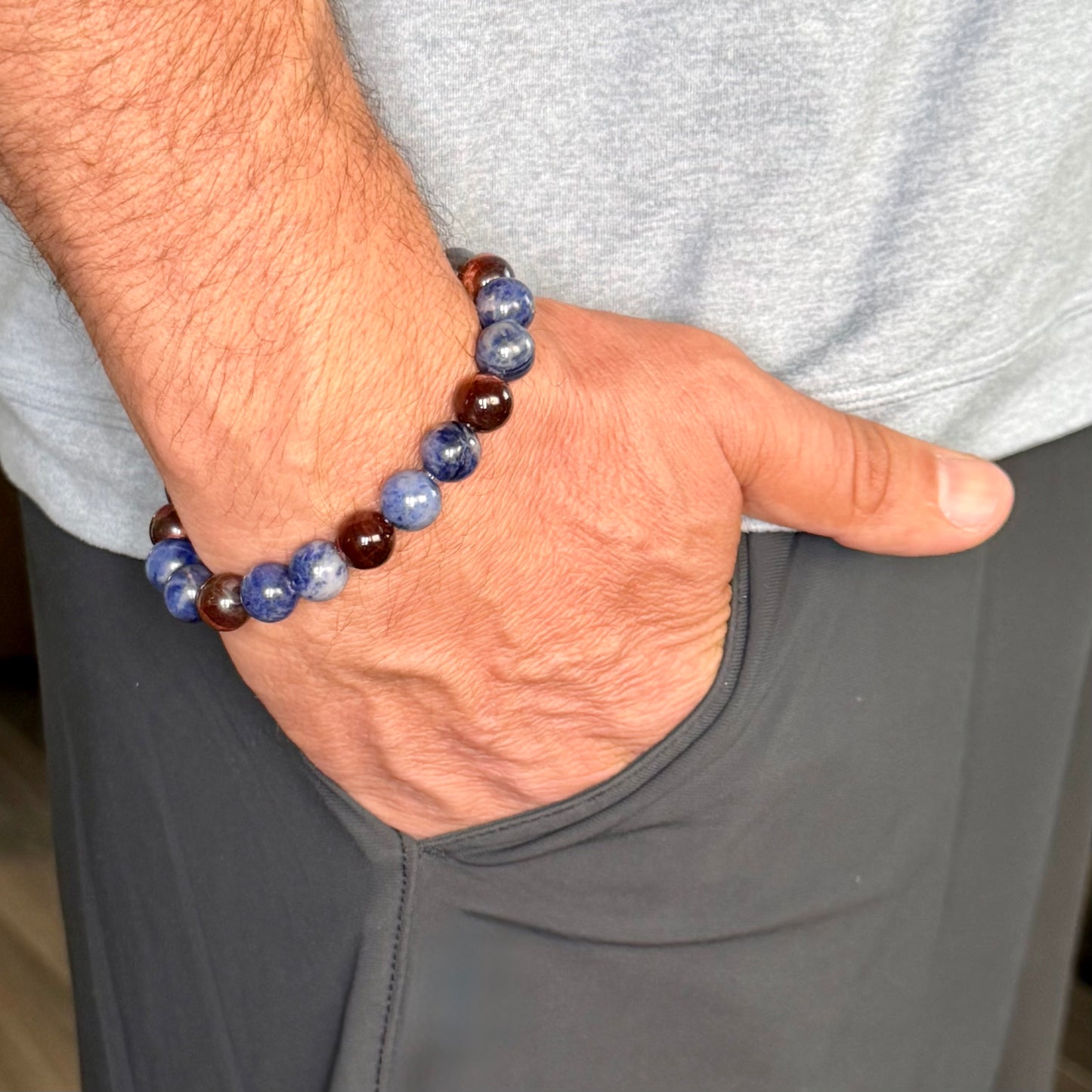A close-up of a man's wrist wearing two beaded bracelets, one blue and white marbled and the other dark brown and blue, paired with a gray t-shirt and pants.