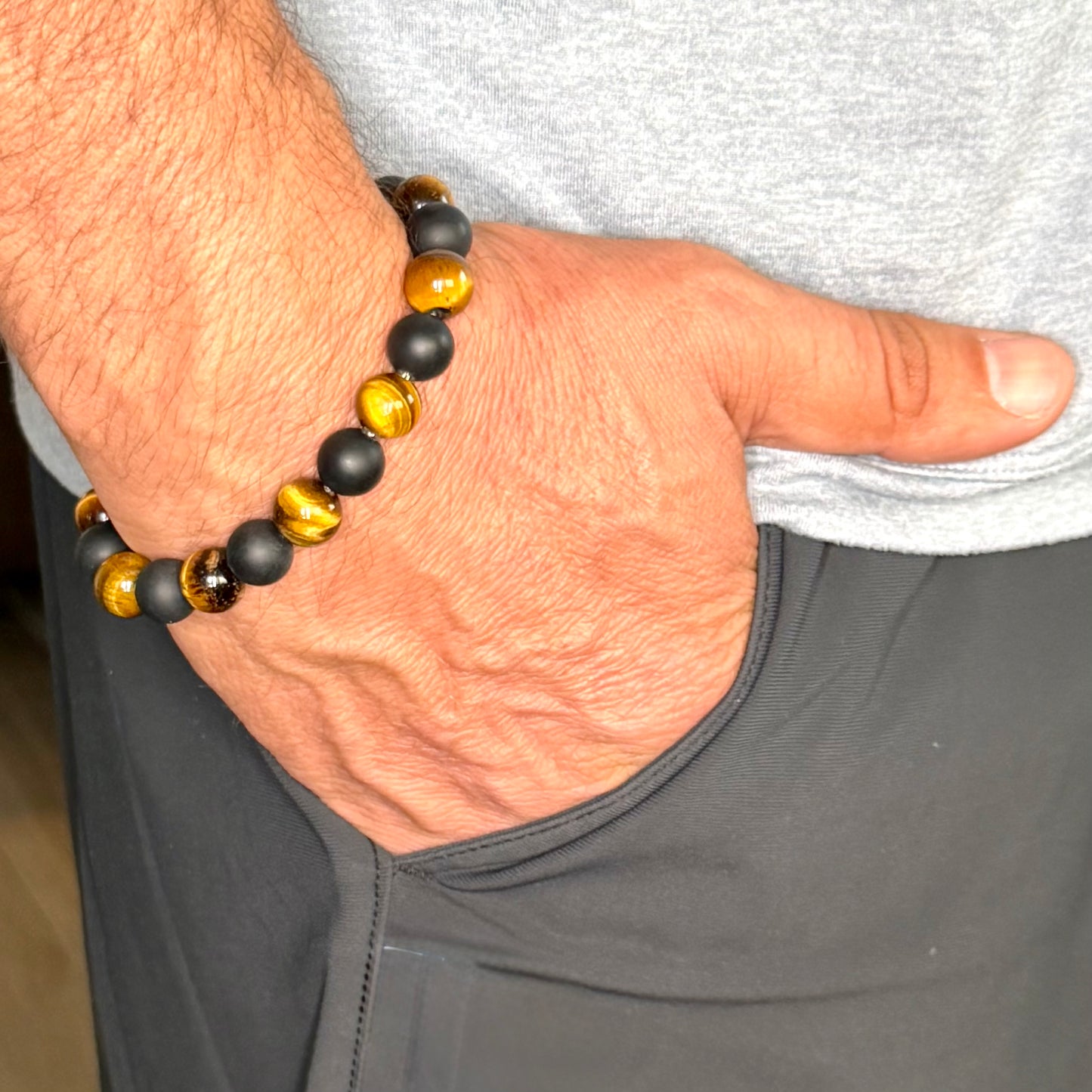 A close-up of a person's hand and wrist wearing a beaded bracelet with alternating matte black and polished golden-brown tiger's eye beads, paired with a gray t-shirt and dark gray athletic pants.