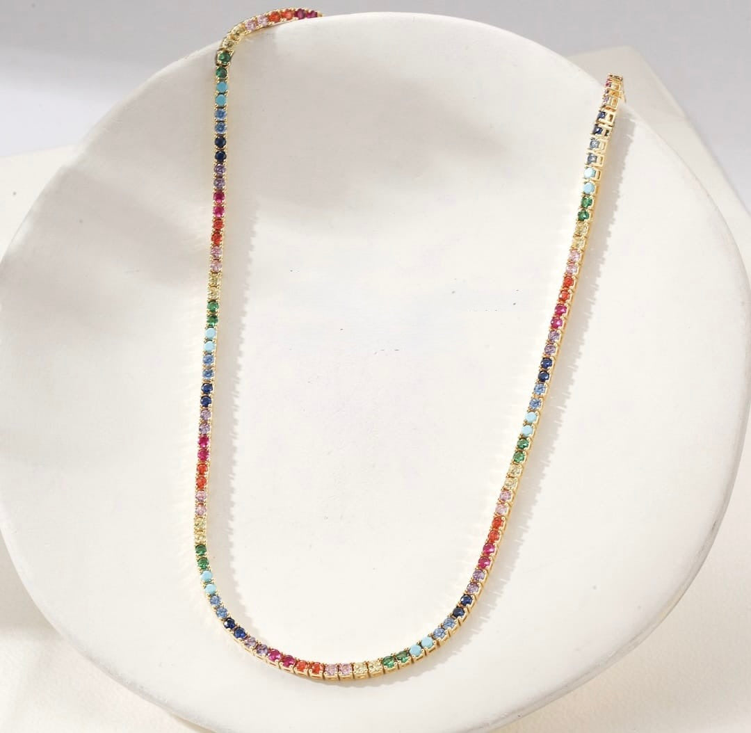 A delicate gold tennis necklace adorned with small, multicolored gemstones, displayed on a white jewelry bust.