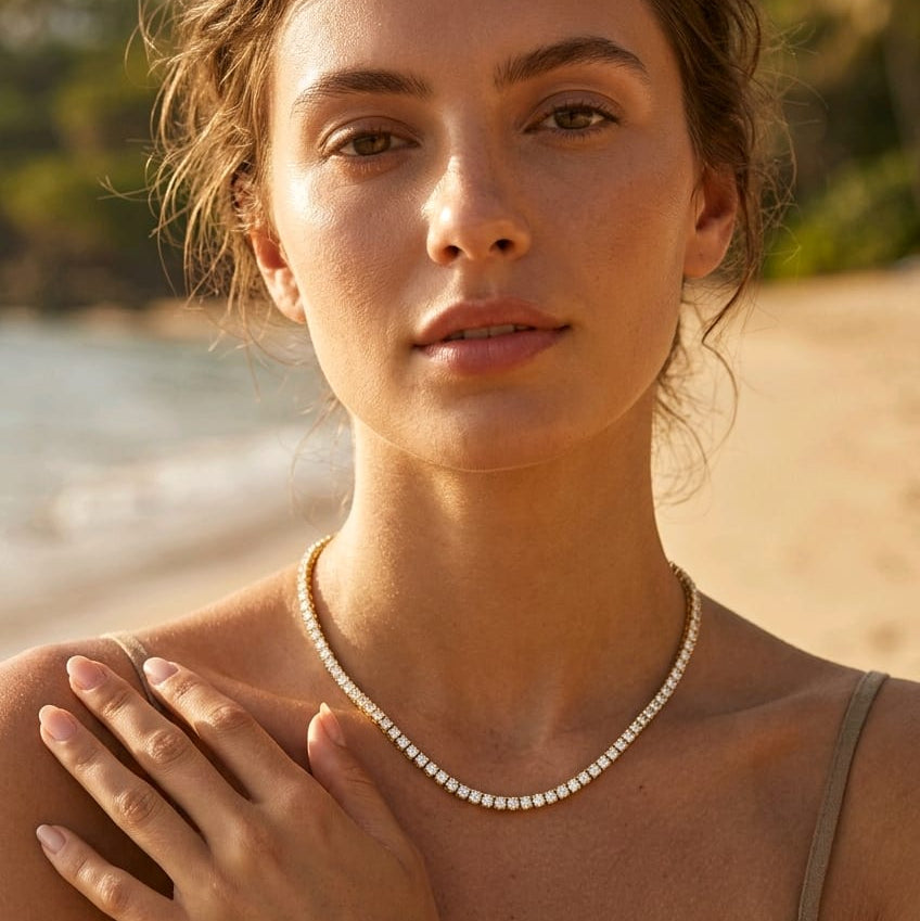 A woman on a beach wearing a delicate white pearl necklace, with her hand resting on her chest. The background shows a soft-focus sandy beach and ocean.