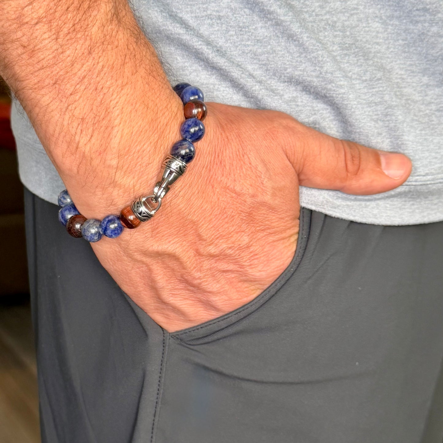 A close-up of a man's hand in his pocket wearing a beaded bracelet. The bracelet features blue and brown translucent beads, a silver-toned metal connector, and a central silver-toned bead with a small red accent.