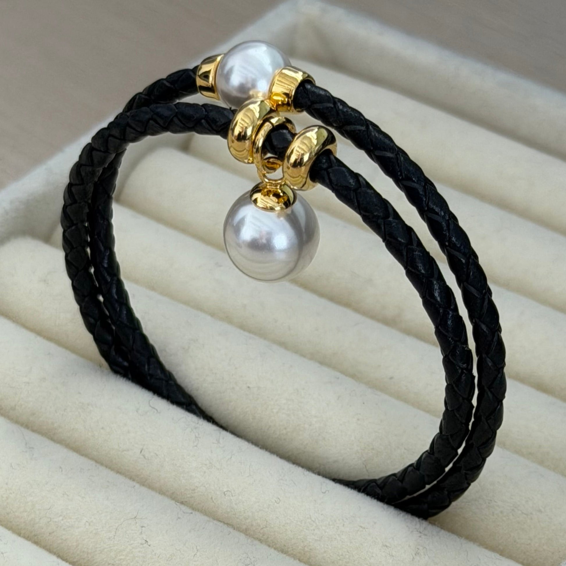 Black cord bracelet with pearl and gold charm on a beige ring holder.