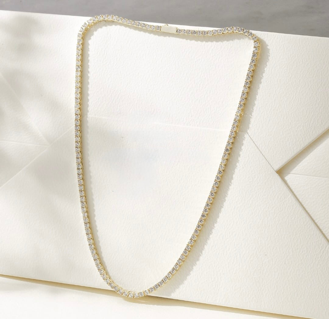 A tennis-style necklace with clear stones set in a yellow-gold metal, arranged in a U-shape on a textured white envelope.