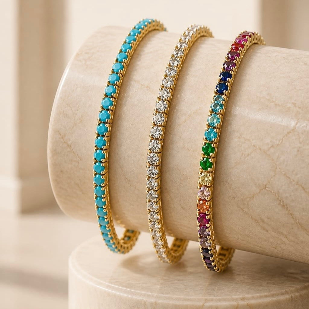 Three gold bangle bracelets displayed on a cream-colored marble stand. One bracelet is set with turquoise-blue stones, one with clear round stones, and one with a multicolor sequence of blue, green, red, orange, and purple stones.