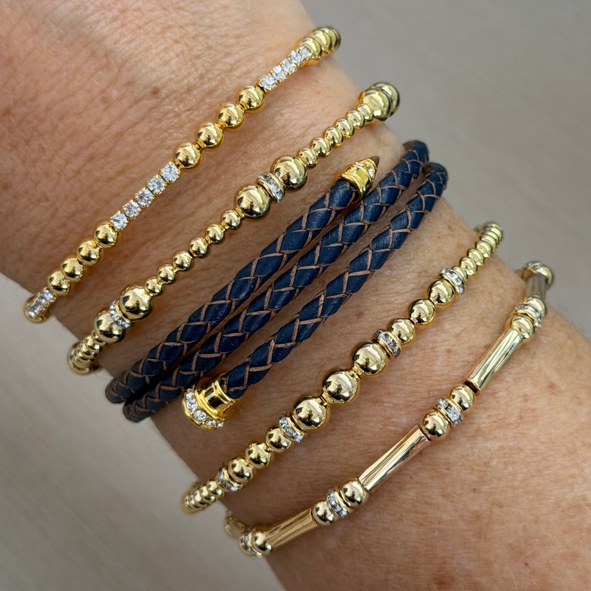 Close-up of a wrist wearing multiple gold and navy bracelets on a neutral background