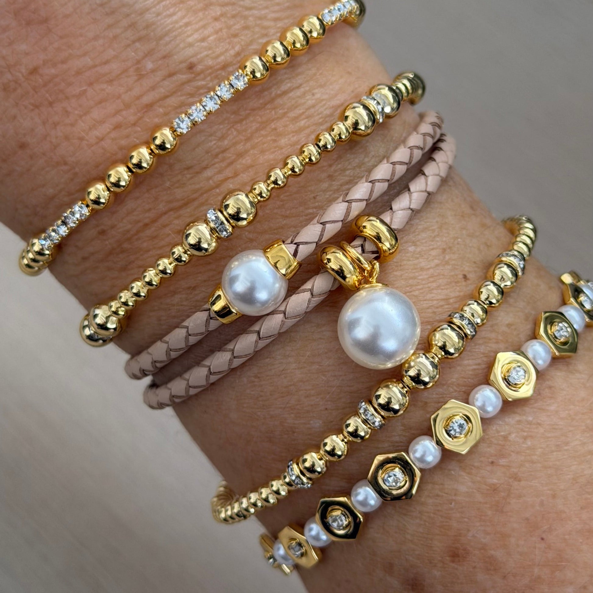 Multiple bracelets on a wrist including gold, pearl, and leather straps.