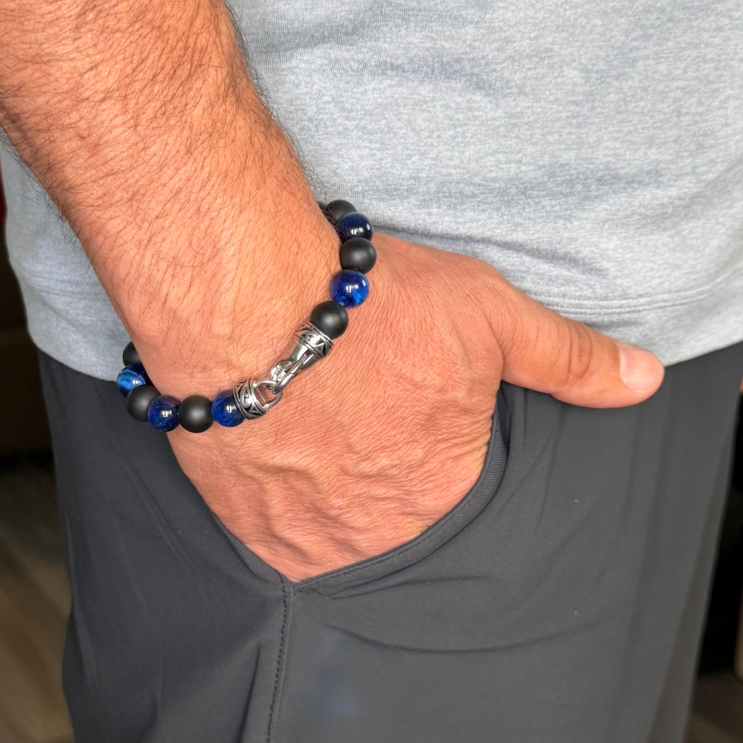 A close-up of a man's hand resting in his pocket, wearing a beaded bracelet with matte black and glossy blue beads, and a silver-toned elephant charm.